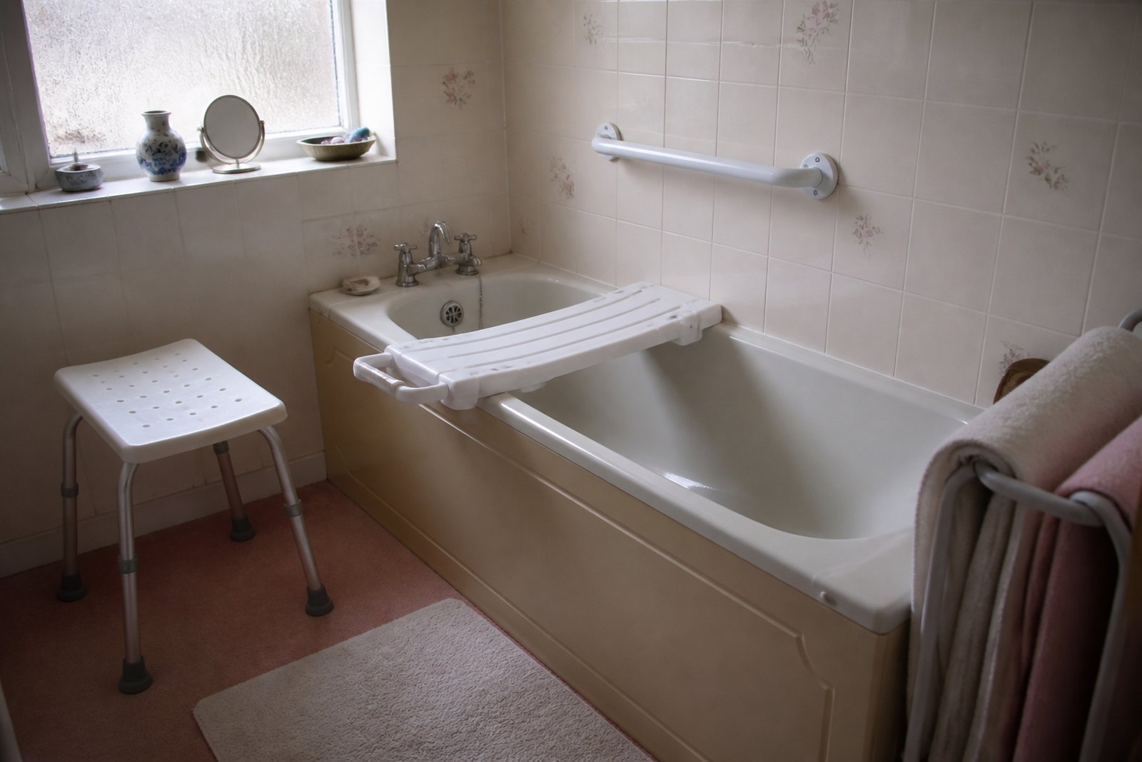 A domestic bathroom with a bath board across the bath, a shower stool beside it and a wall-fixed grab rail