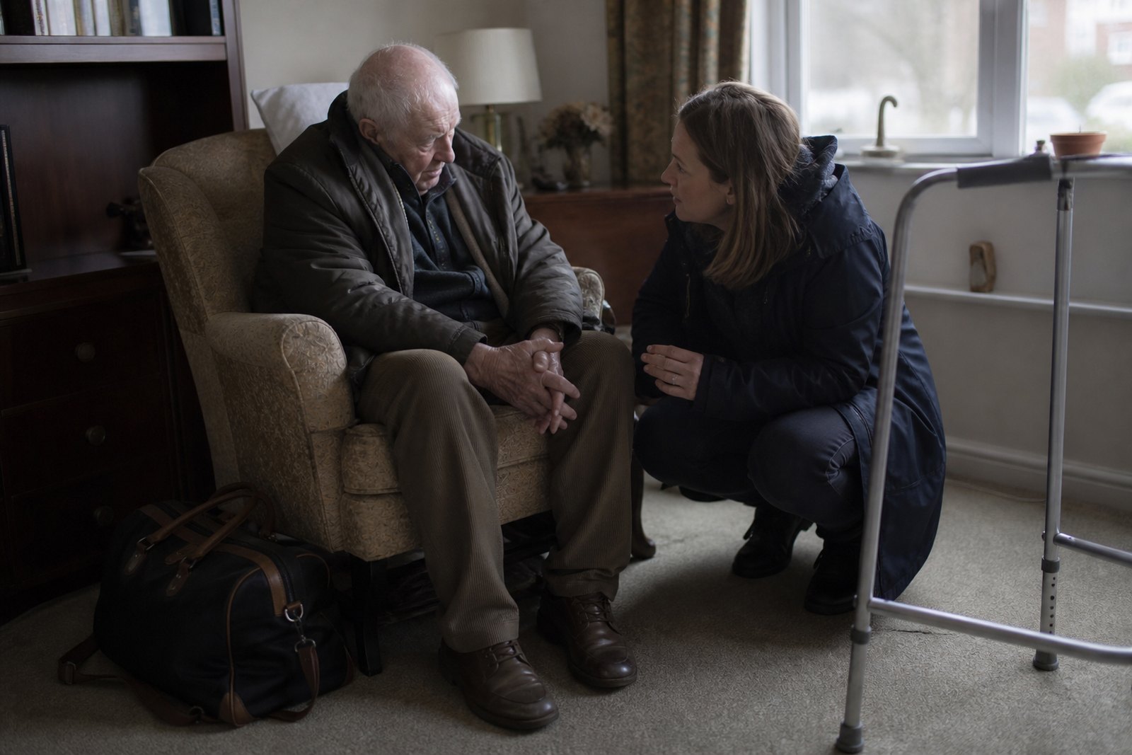 An older man newly home from hospital sitting in his armchair while his daughter talks with him