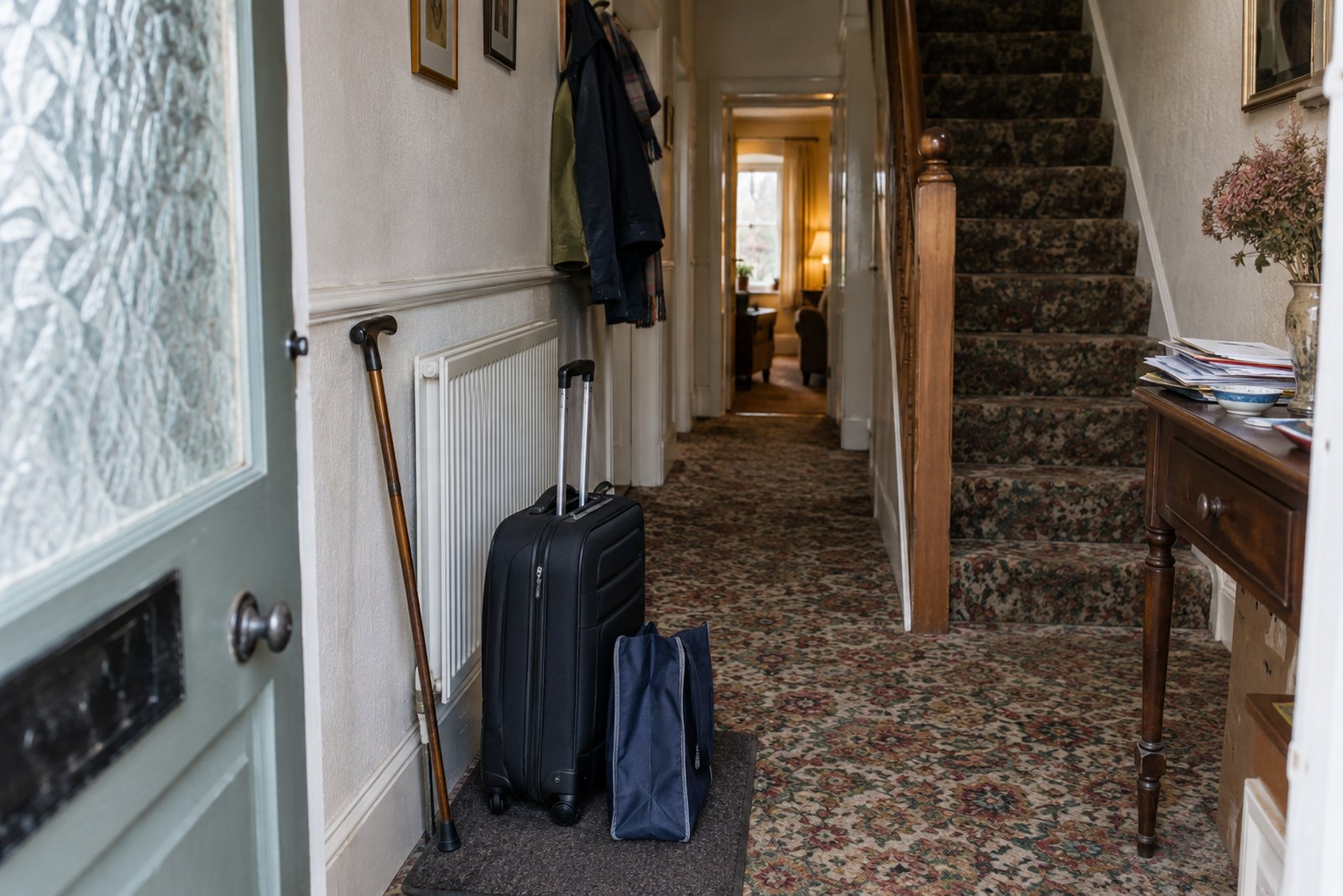 A front hallway with a small suitcase set down on the mat and a walking stick leaning by the radiator, with warm light from a room further down the hall