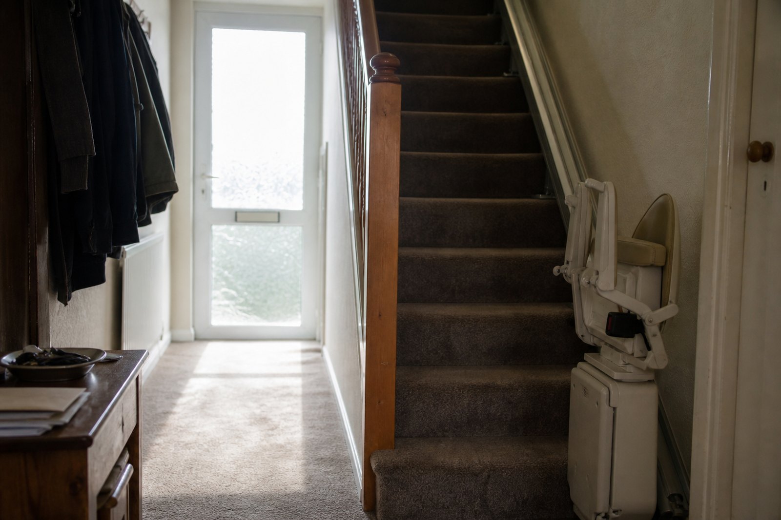A stairlift with its seat folded at the bottom of a carpeted staircase in a domestic hallway