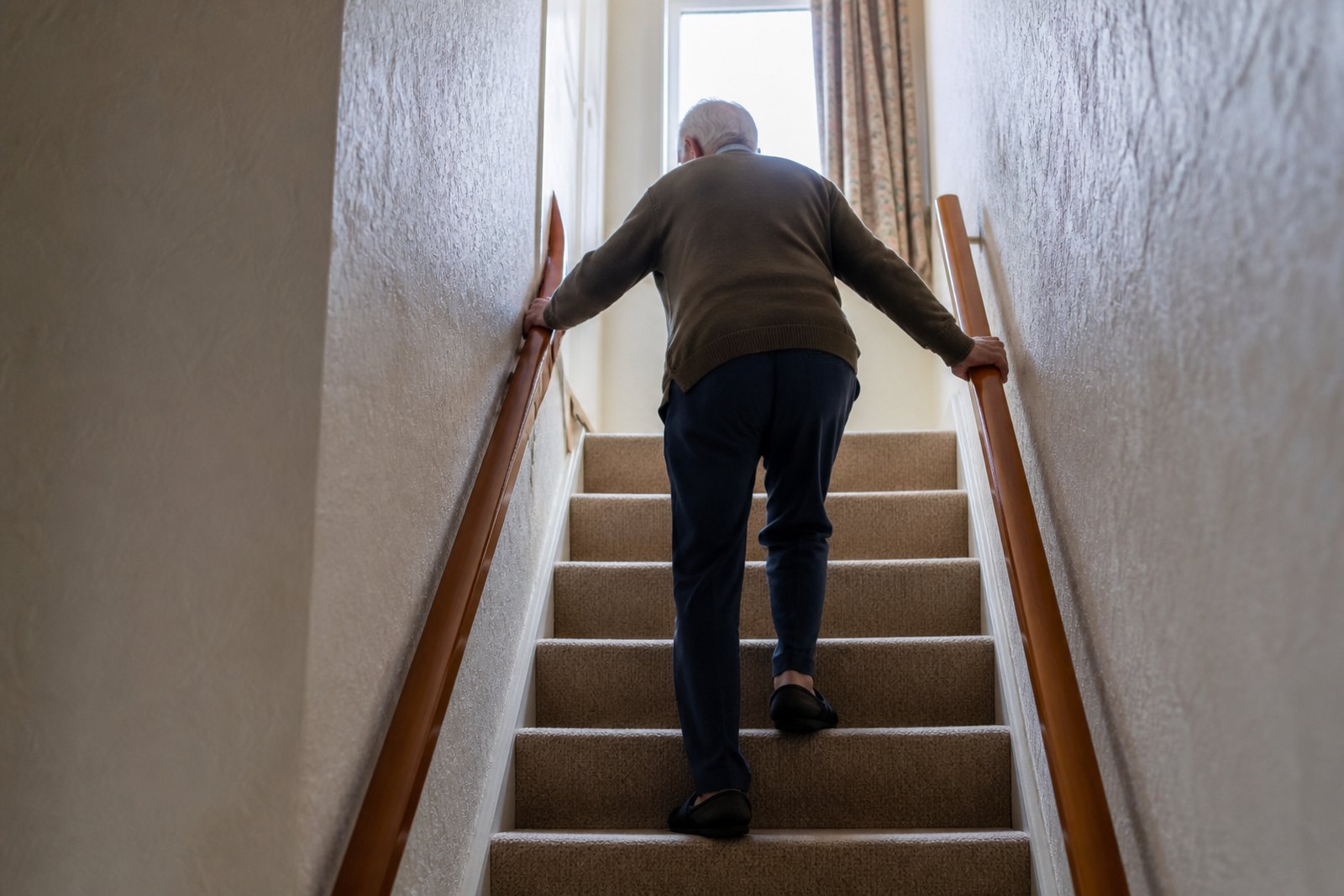 An older man seen from behind climbing a staircase using handrails on both sides