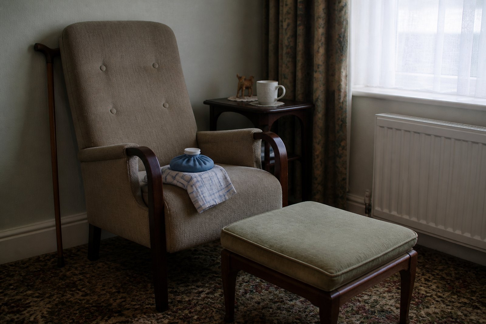 A high firm armchair by a window with a cushioned footstool in front of it and an ice pack wrapped in a tea towel over the arm