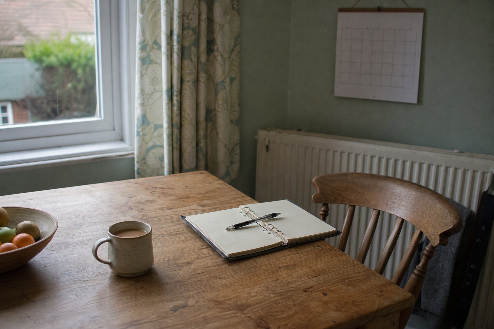 An open ring binder of care notes with a pen and a mug of tea on a kitchen table, a blank wall calendar behind