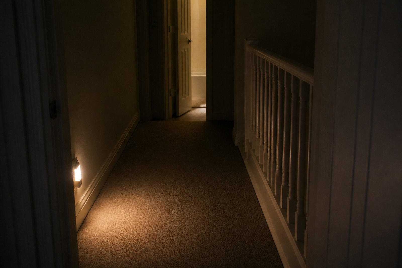 A dark landing at night lit by a plug-in night light, with a clear floor and the bathroom door ajar