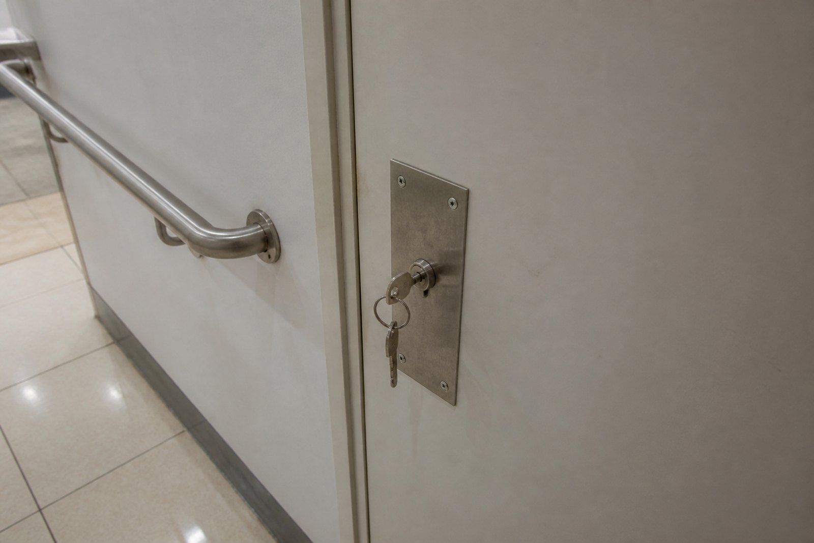 A chunky key in the lock of a closed accessible toilet door in a public building, with a handrail on the wall beside it
