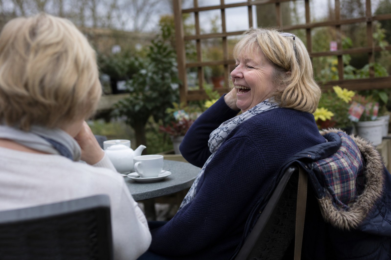 Two women laughing over a pot of tea at an outdoor cafe table
