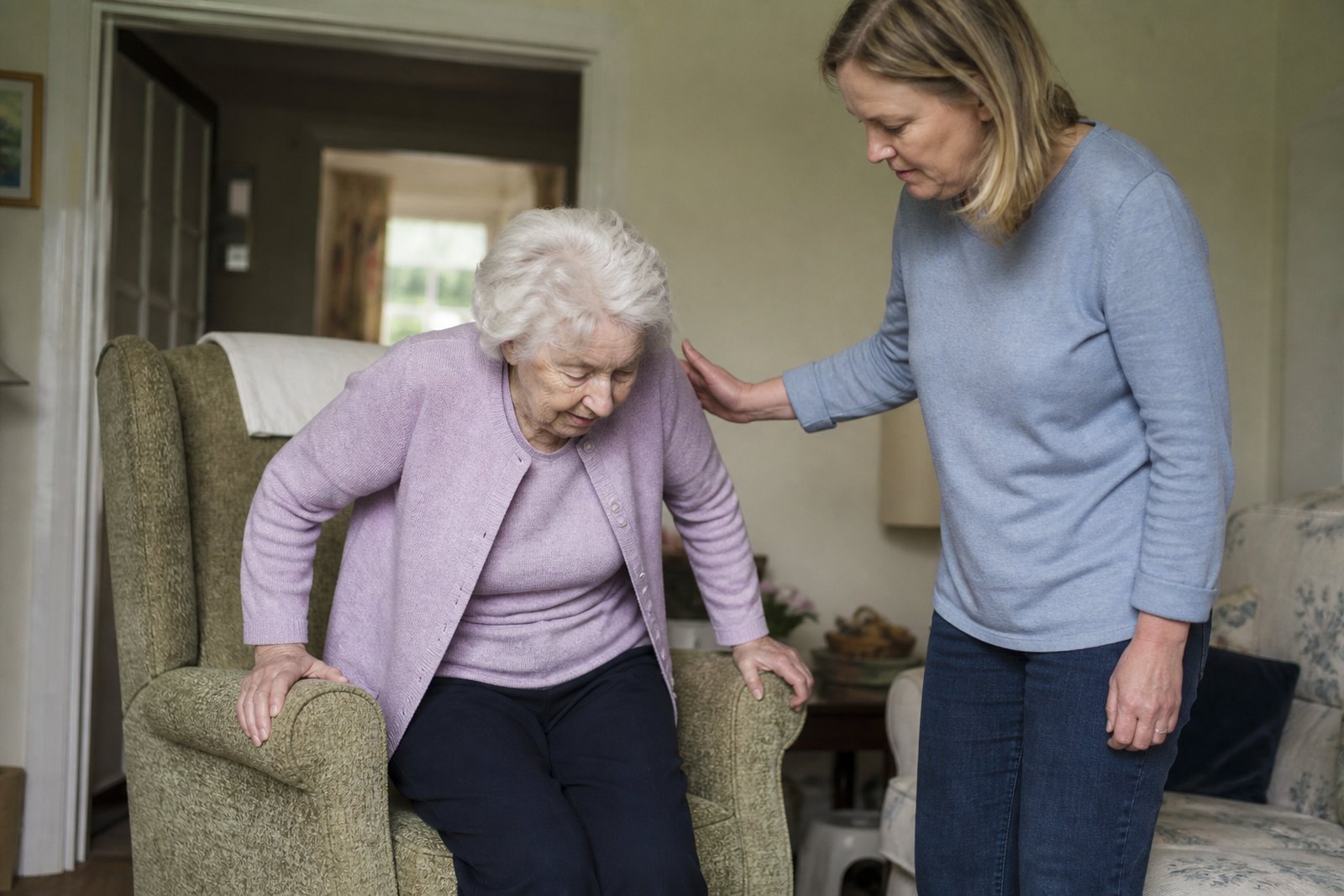 An older woman pushing up from her armchair while her daughter stands beside her, steadying her back